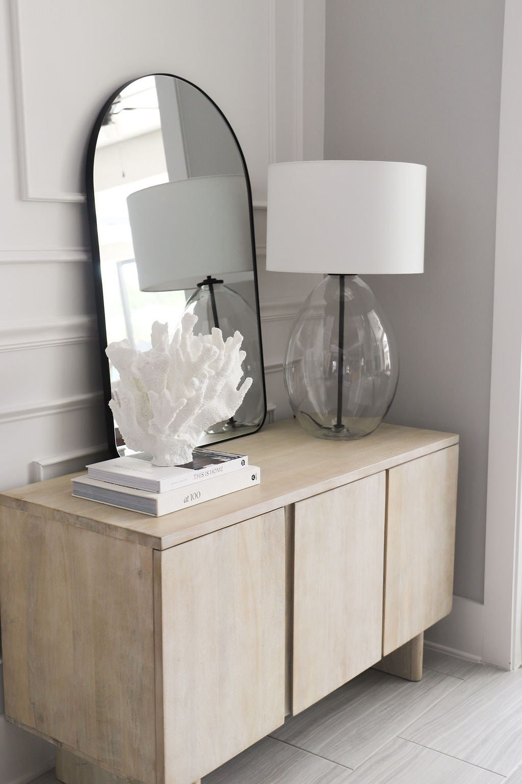 A light wood cabinet that features a mirror with a black rim, a glass lamp with white shade, a stack of books and a coral statue.