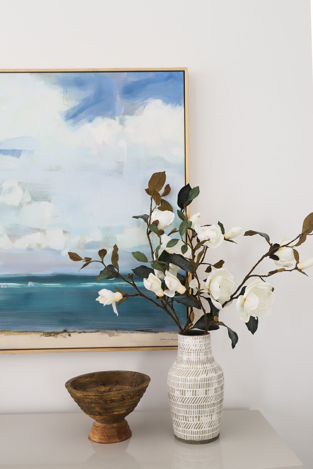 A ceramic vase of magnolia stems and a wooden bowl sit on top a shelf. A painting of the ocean is hung behind the items.