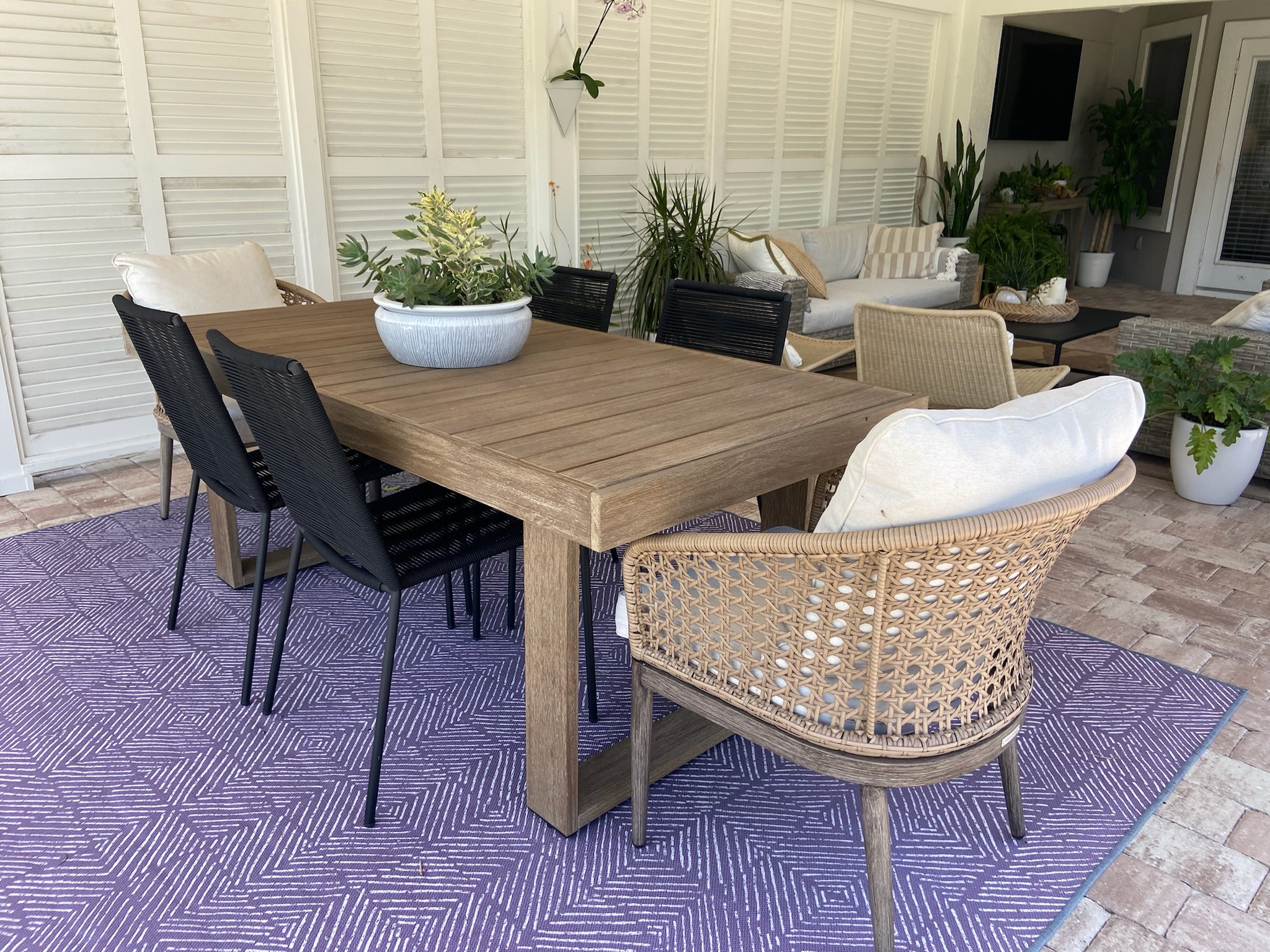 This gorgeous patio space is the perfect spot to sit and enjoy the outdoors. A large wooden table and a mixture of chairs sits on top a colorful rug. 