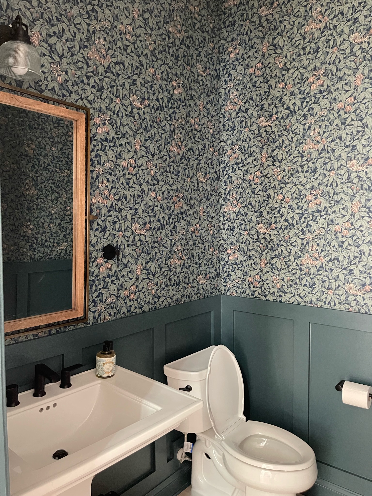 This bathroom has wood paneling halfway up the walls that is painted a dark blue . The other half of the wall uses a printed wallpaper that matches the blue wall below. 