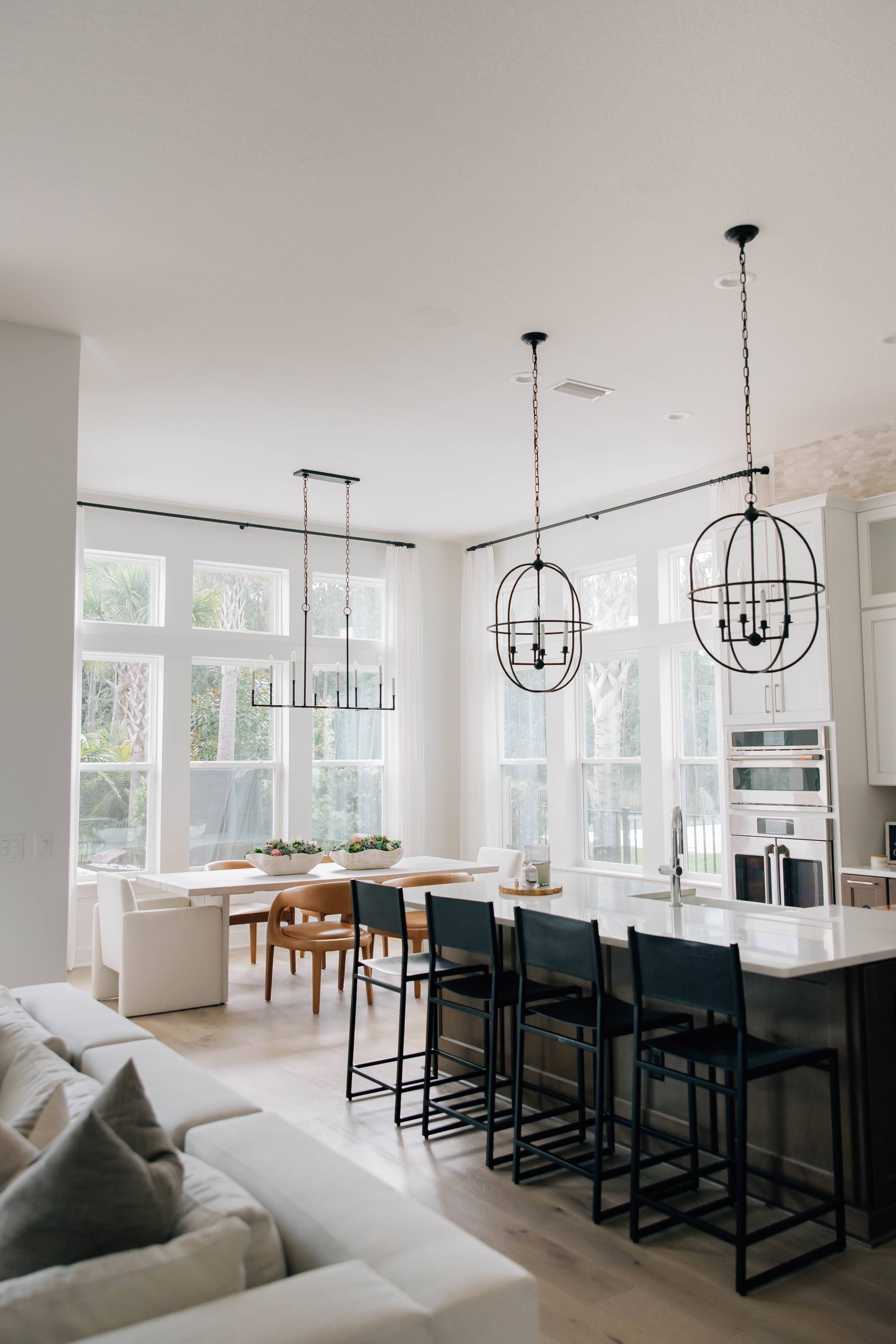 A modern, open kitchen and dining area exemplifies stunning interior design by Welcome Home Styling with white walls, high ceilings, and large windows. Recent renovation highlights include a central island with bar stools, a dining table with chairs, and three pendant lights, all beautifully illuminated by natural light.