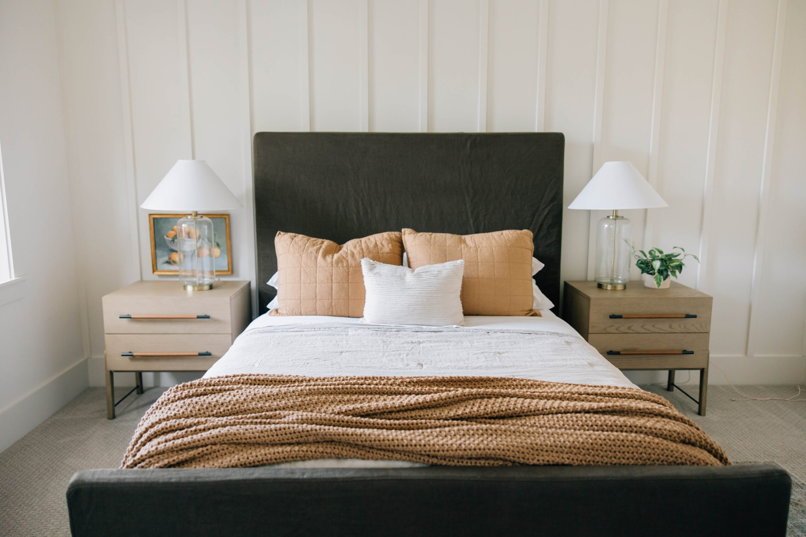 A neatly made bed with a dark headboard features three pillows and a brown knitted throw. Two wooden nightstands with white lamps and a small plant are on each side. Designed by a Jacksonville, Florida interior designer Jessica Woodward and the Welcome Home Styling team, the room's vertical paneling creates a cozy, modern atmosphere.