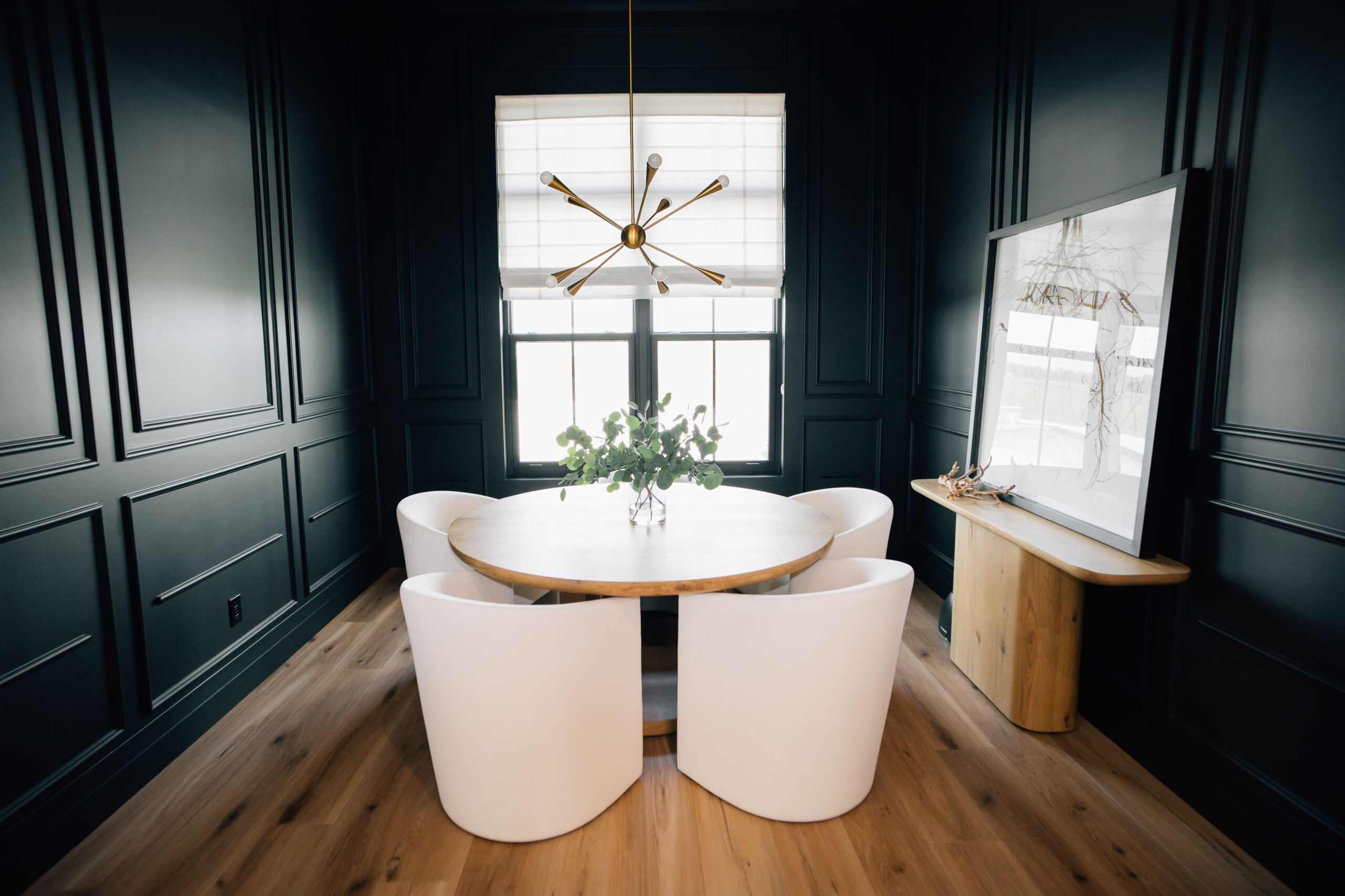 Moody and Modern Dining Room The dining room with dark wainscoting on the walls, a natural wood round table, and sculptural dining chairs designed by Welcome Home Styling.