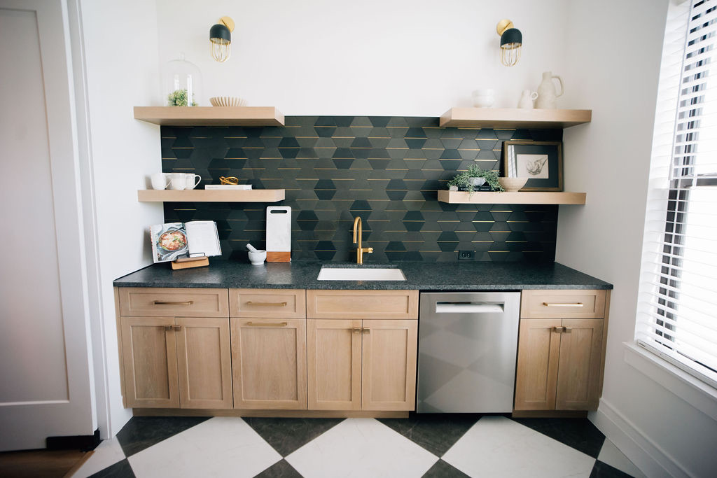 Butler’s pantry with light wood cabinets, floating shelves, black geometric tile backsplash, and gold faucet designed as part of a Ponte Vedra interior design project.