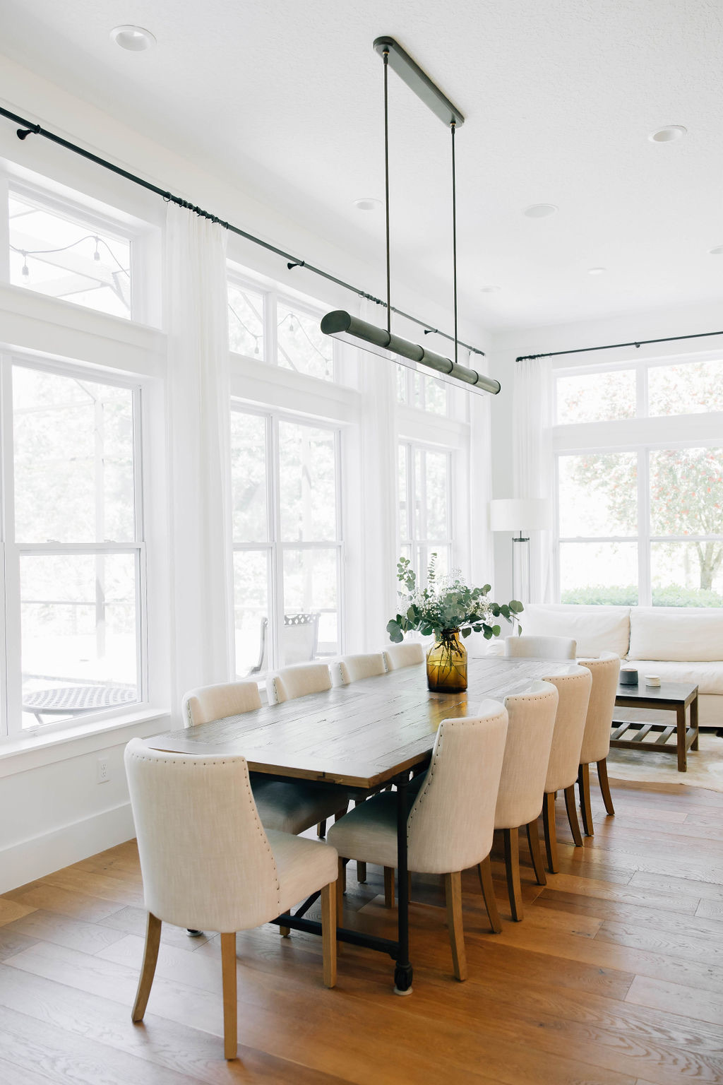 Dining room with long wood table, cream upholstered chairs, modern black light fixture, and floor-to-ceiling windows as part of a Ponte Vedra interior design project.