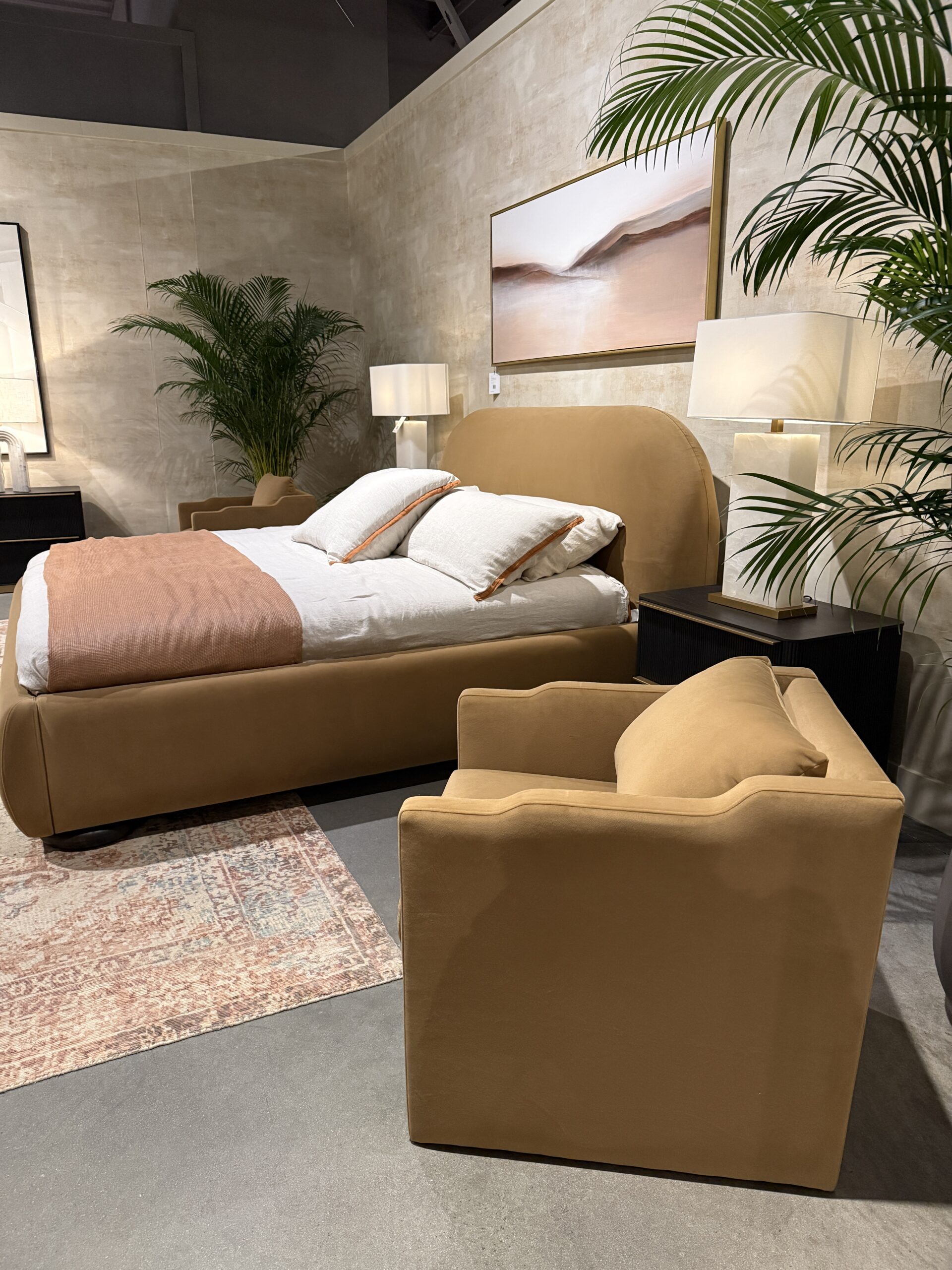 Light brown velvet bed and matching chair in a modern showroom, styled with neutral finishes and greenery.