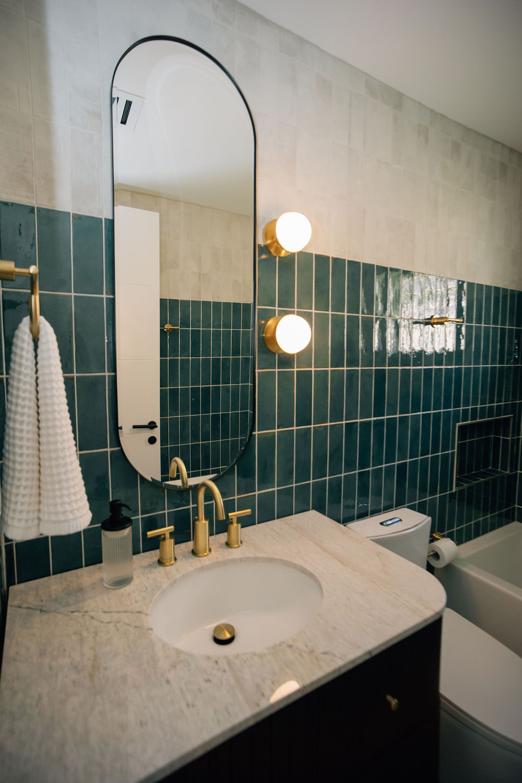Bathroom with oval mirror, unique circular lights, green tile, and gold accents.