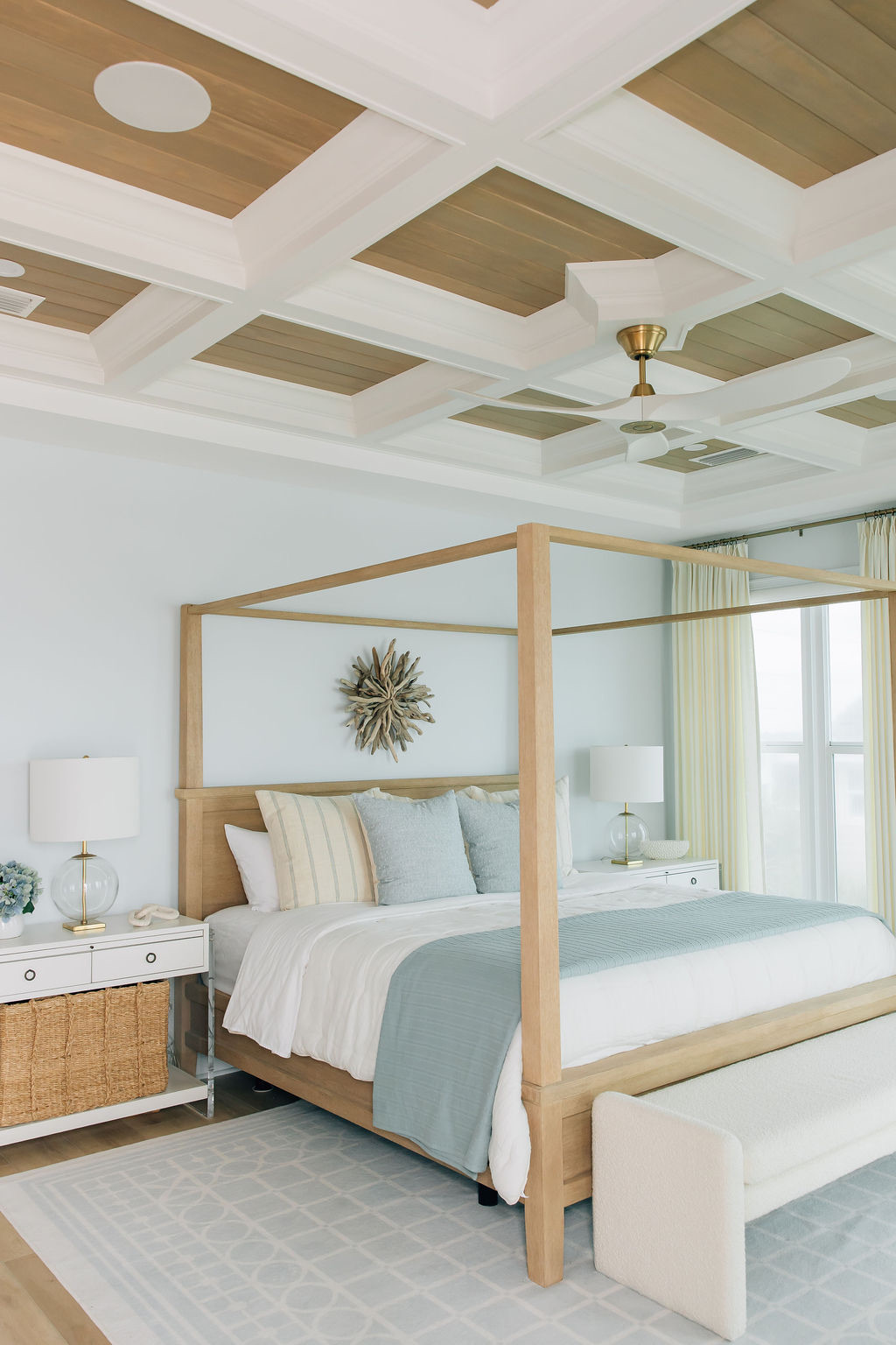Bedroom with a light wood four-poster bed, tray ceiling with light wood accents, white bedside tables with wicker storage bins, and blue and white bed linens. 