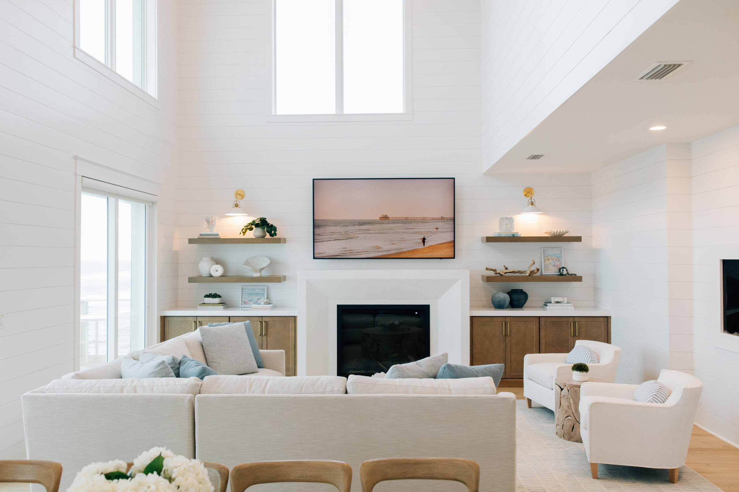 Coastal living room with white seating, a fireplace, floating shelves, tall windows, and soft blue accents throughout the space.