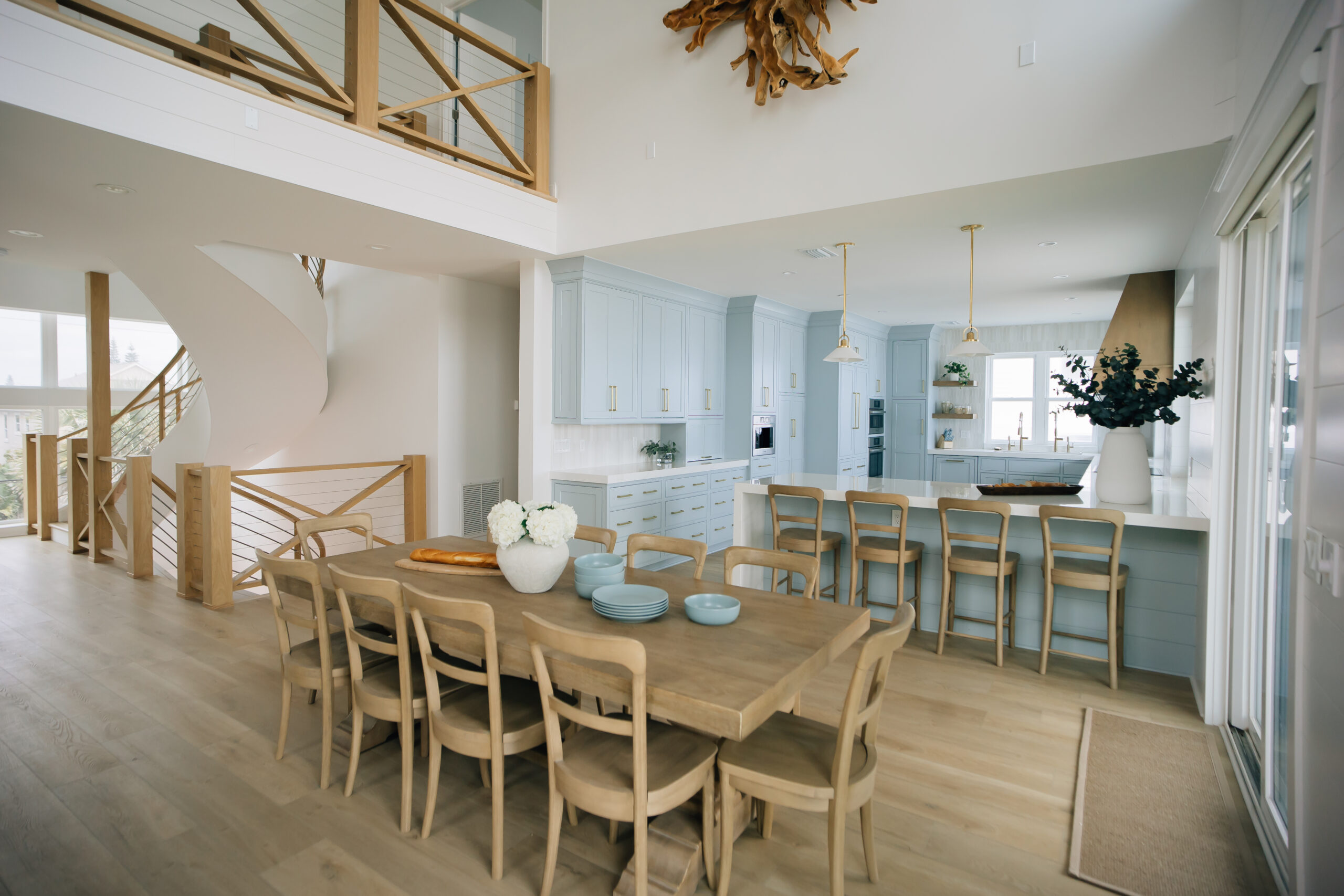 Light blue coastal kitchen with a large dining table, soaring ceilings, natural wood details, and expansive windows that fill the space with sunlight.