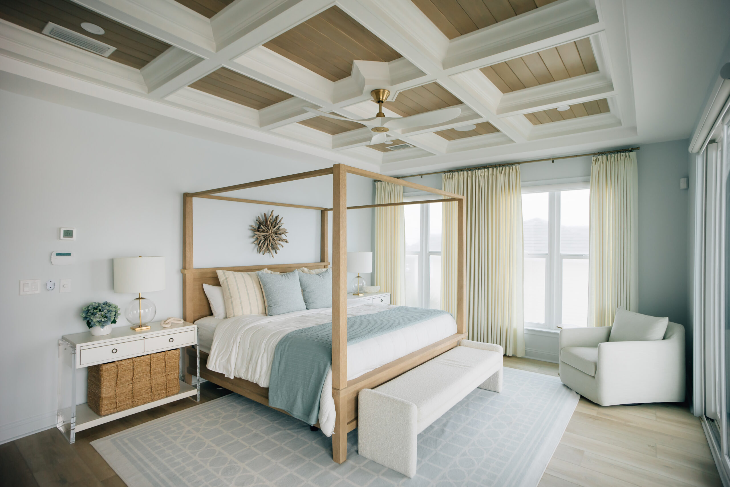 Light-filled bedroom with a wood canopy bed, upholstered bench, coffered ceiling, soft blue bedding, and floor-to-ceiling drapery.