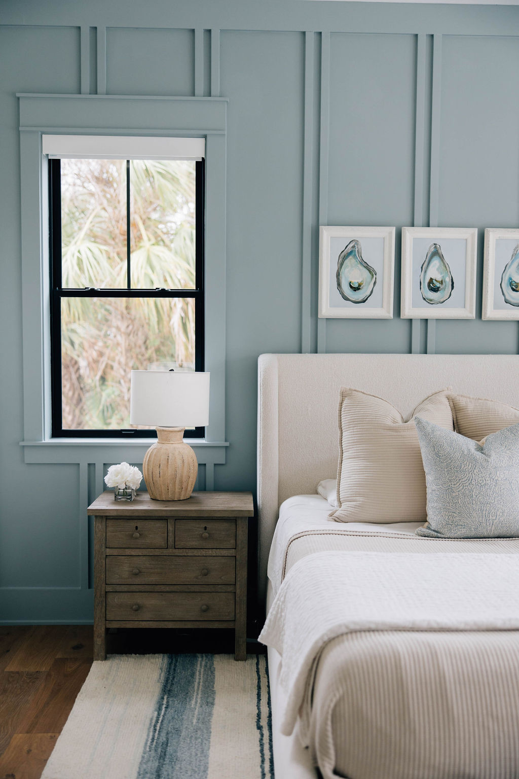 Bedroom corner with a light upholstered headboard, blue accent wall, framed coastal artwork, wood nightstand, and decorative pillows.