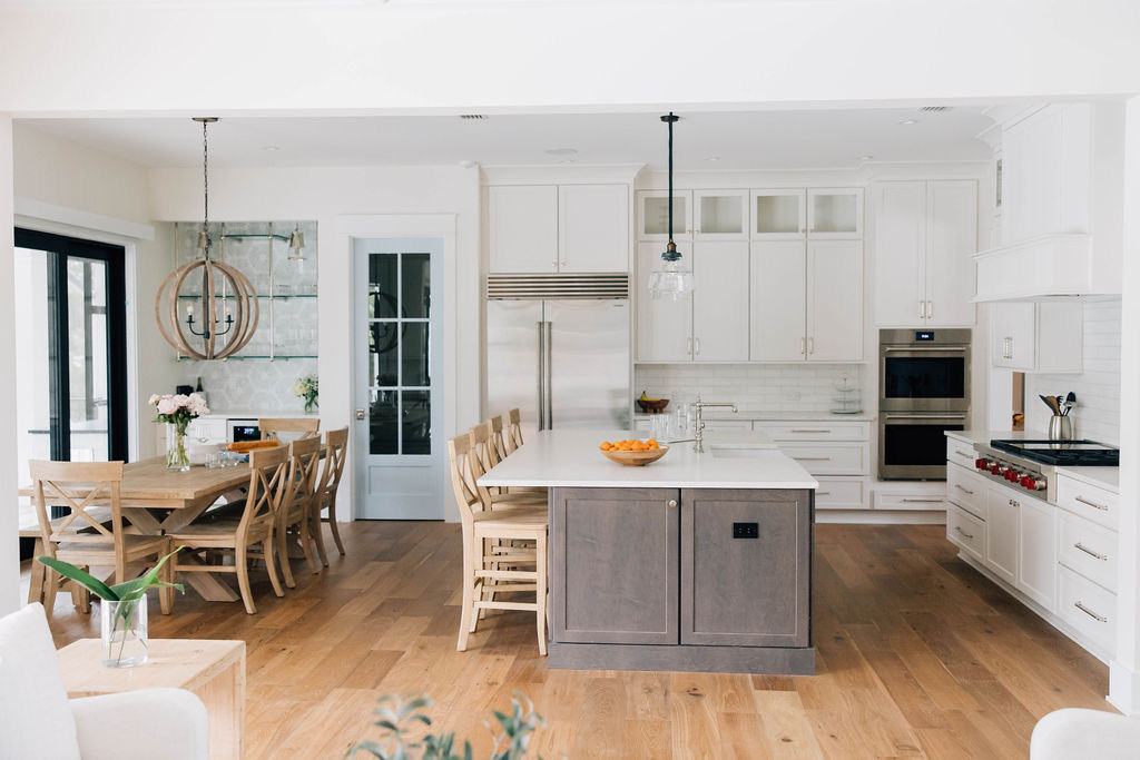 White coastal kitchen with a large island, wood flooring, bar seating, and an adjacent dining table in an open-concept layout.