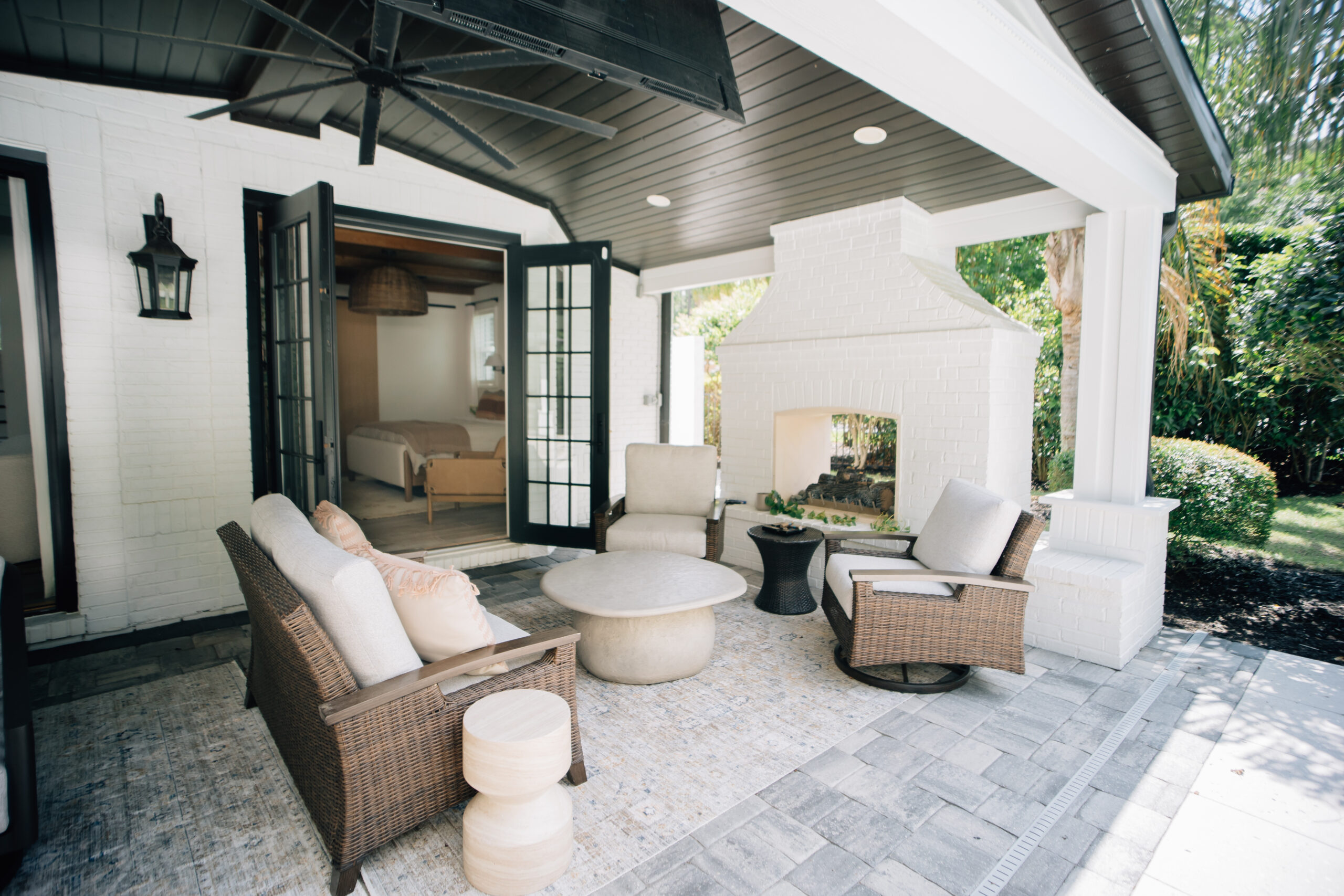 A covered outdoor patio with wicker seating, cushioned chairs, a white brick fireplace, ceiling fan, and views of a landscaped backyard.