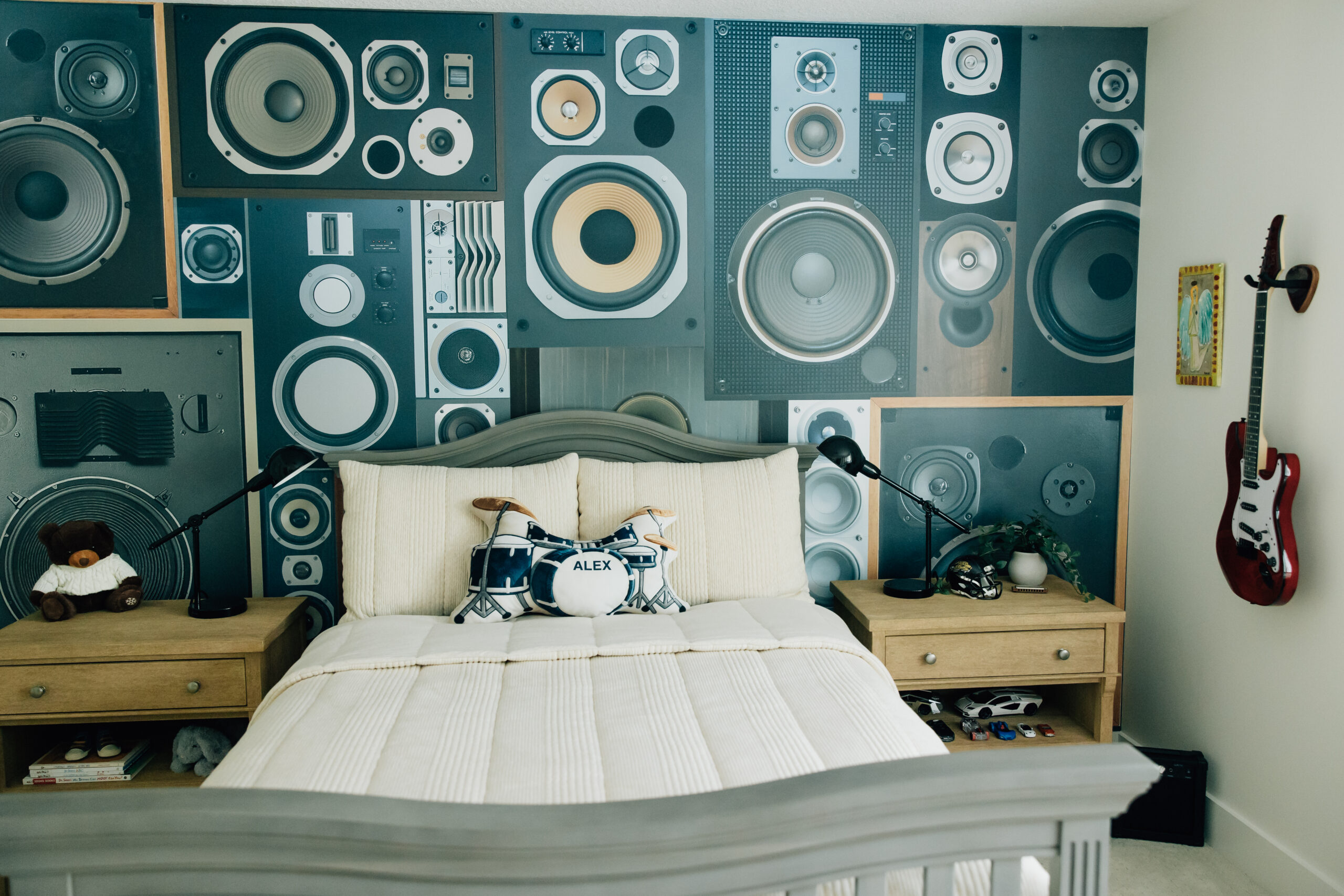 A child's bedroom featuring a speaker-themed accent wall, light wood furniture, a gray bed frame, music-inspired decor, and personalized drum pillows.