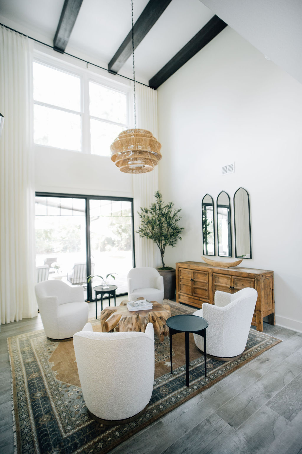 Modern living room with vaulted ceilings, exposed beams, oversized windows, white accent chairs, woven pendant light, and abundant natural light.
