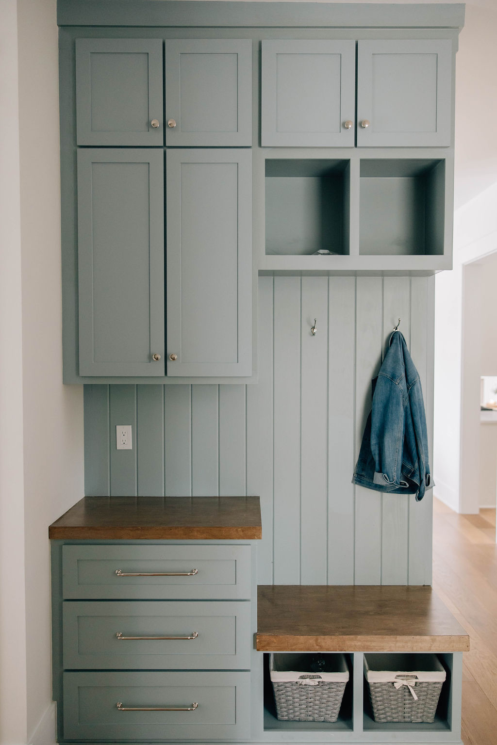 Built-in mudroom with sage green cabinetry, wood bench seating, open cubbies, drawers, baskets, and wall hooks for coats and everyday storage.