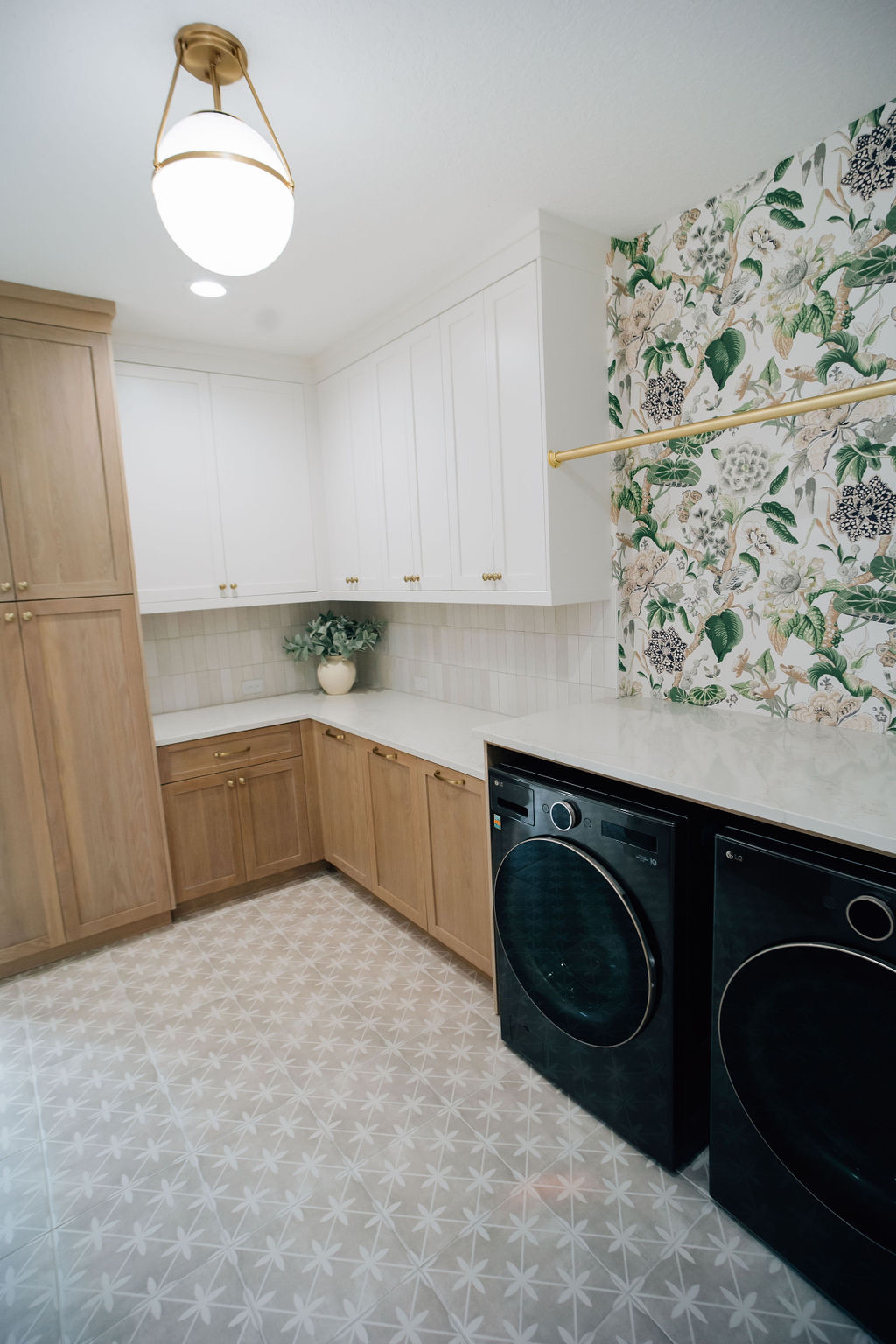 Bright laundry room with white and natural wood cabinets, patterned wallpaper, quartz countertops, black front-loading washer and dryer, and decorative tile flooring.