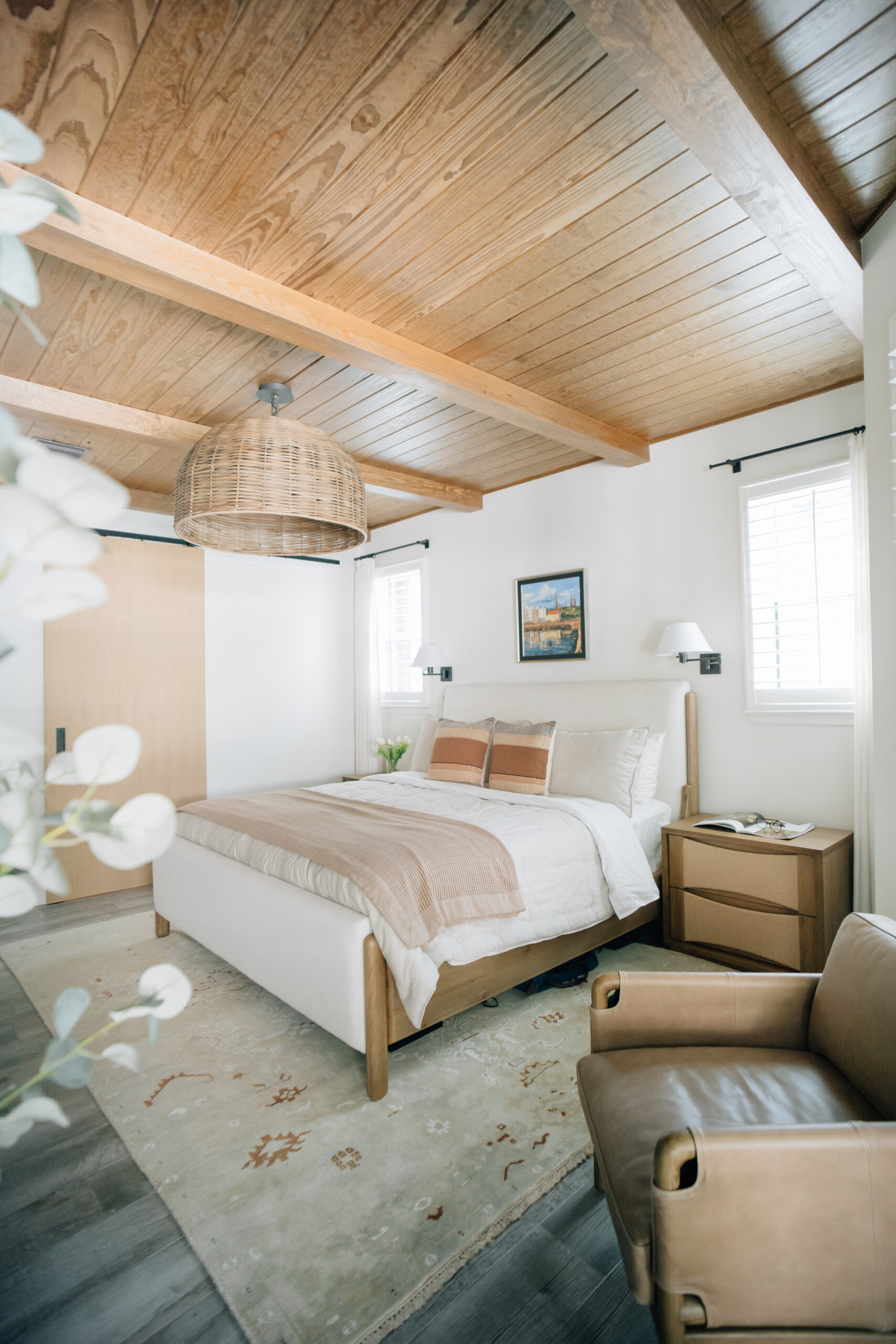 Primary bedroom designed by a new home construction interior designer featuring exposed wood ceiling beams, upholstered bed, neutral furnishings, and warm natural textures.