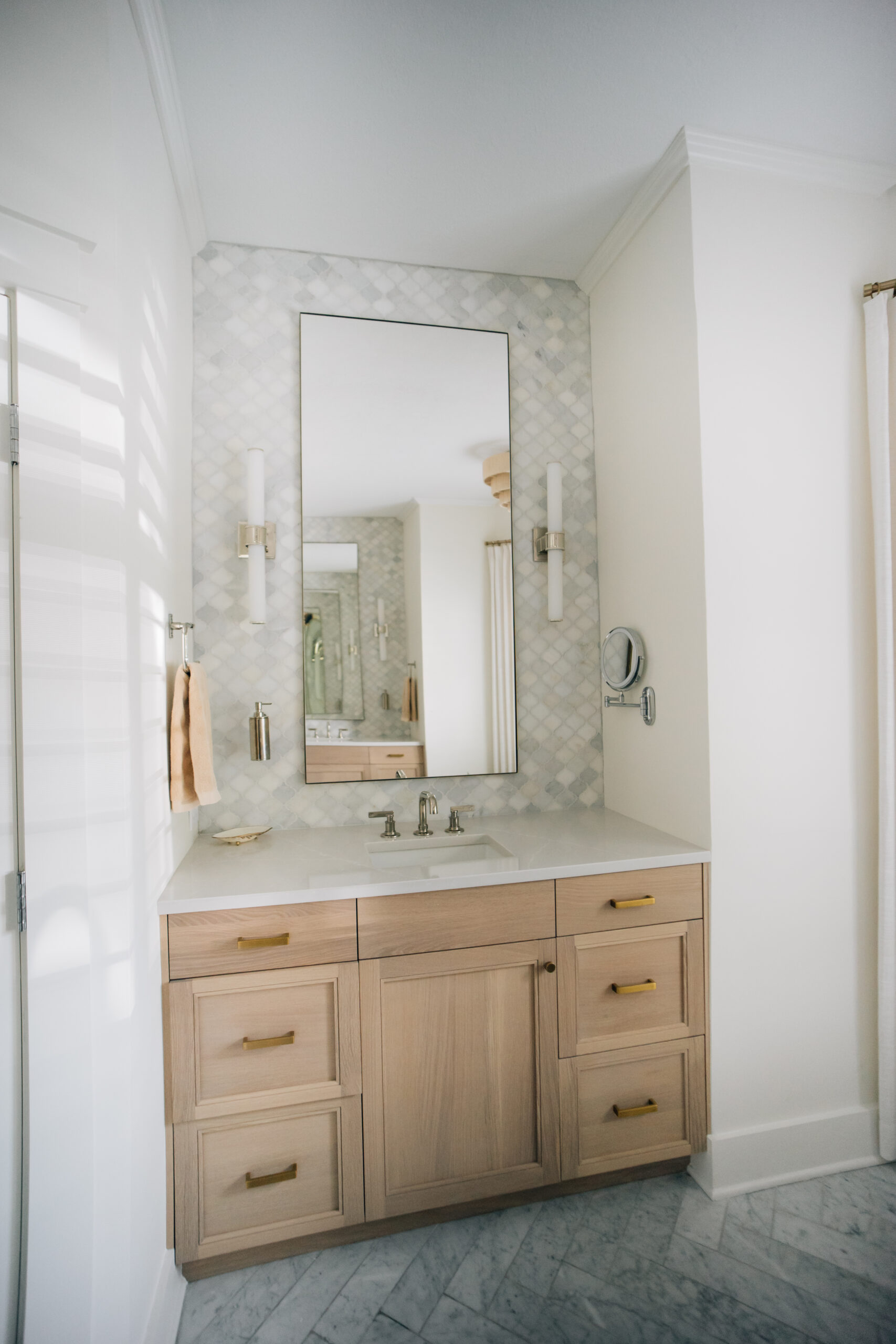 Custom bathroom vanity in a new construction home featuring light wood cabinetry, brass hardware, marble-inspired tile, wall sconces, and a modern framed mirror.