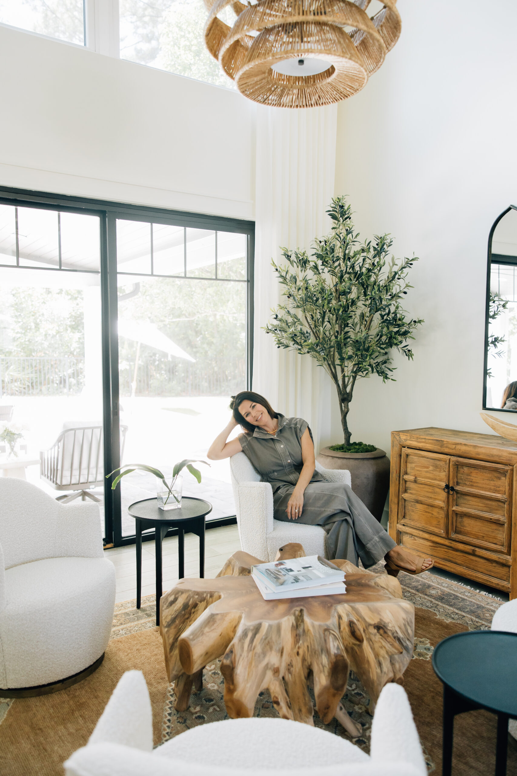 Interior designer seated in a bright modern living room with oversized windows, white accent chairs, a natural wood coffee table, woven pendant light, and indoor greenery.