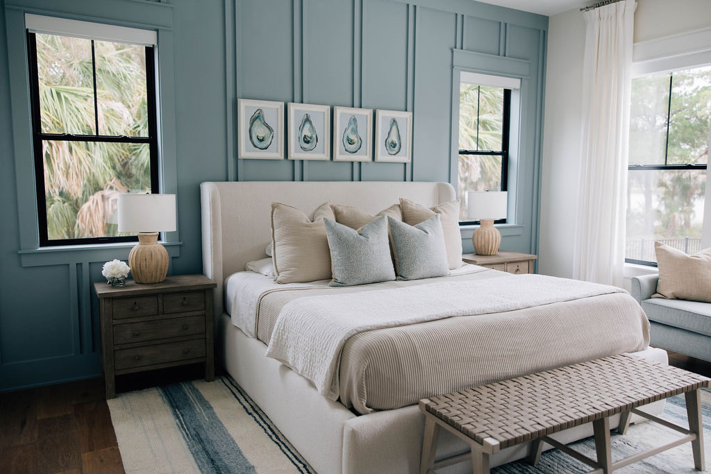Primary bedroom with a blue paneled accent wall, upholstered bed, neutral bedding, wood nightstands, woven bench, and framed artwork.