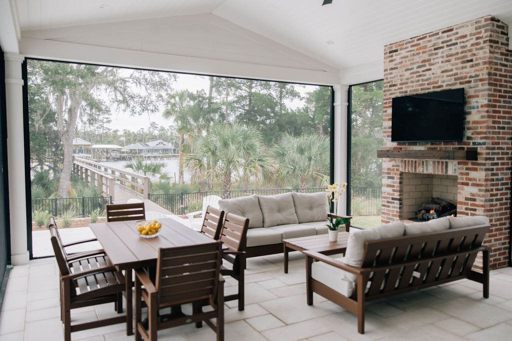 Screened outdoor living area with wood dining furniture, cushioned seating, a brick fireplace, and views of palm trees and water.