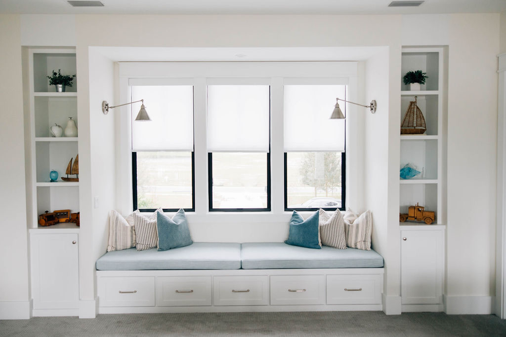 Built-in window seat with blue cushions, storage drawers, white shelving, wall-mounted reading lights, and decorative accessories.