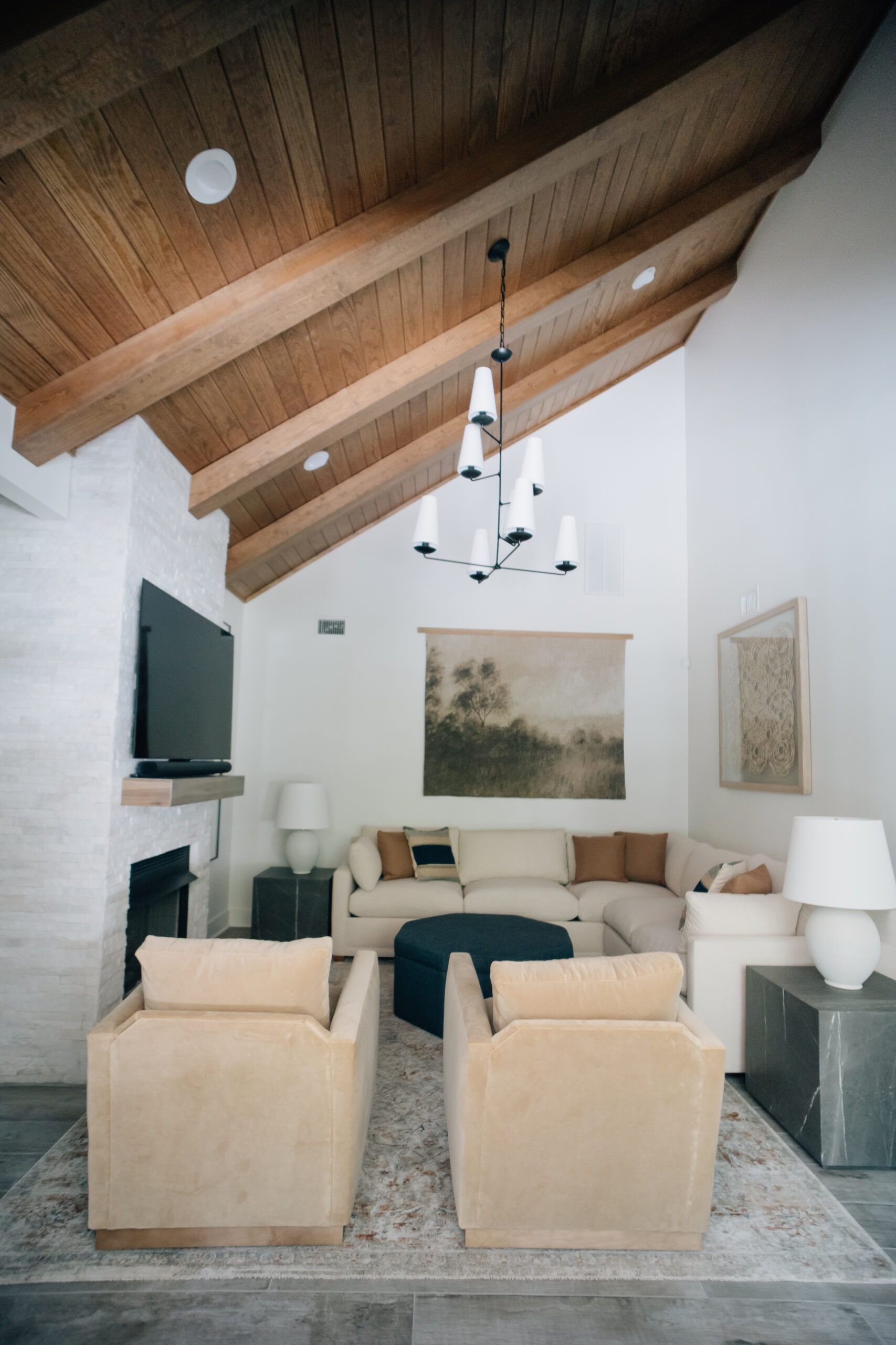 Living room with a vaulted wood ceiling and exposed beams, white sectional, two cream armchairs, black chandelier, and white brick fireplace.