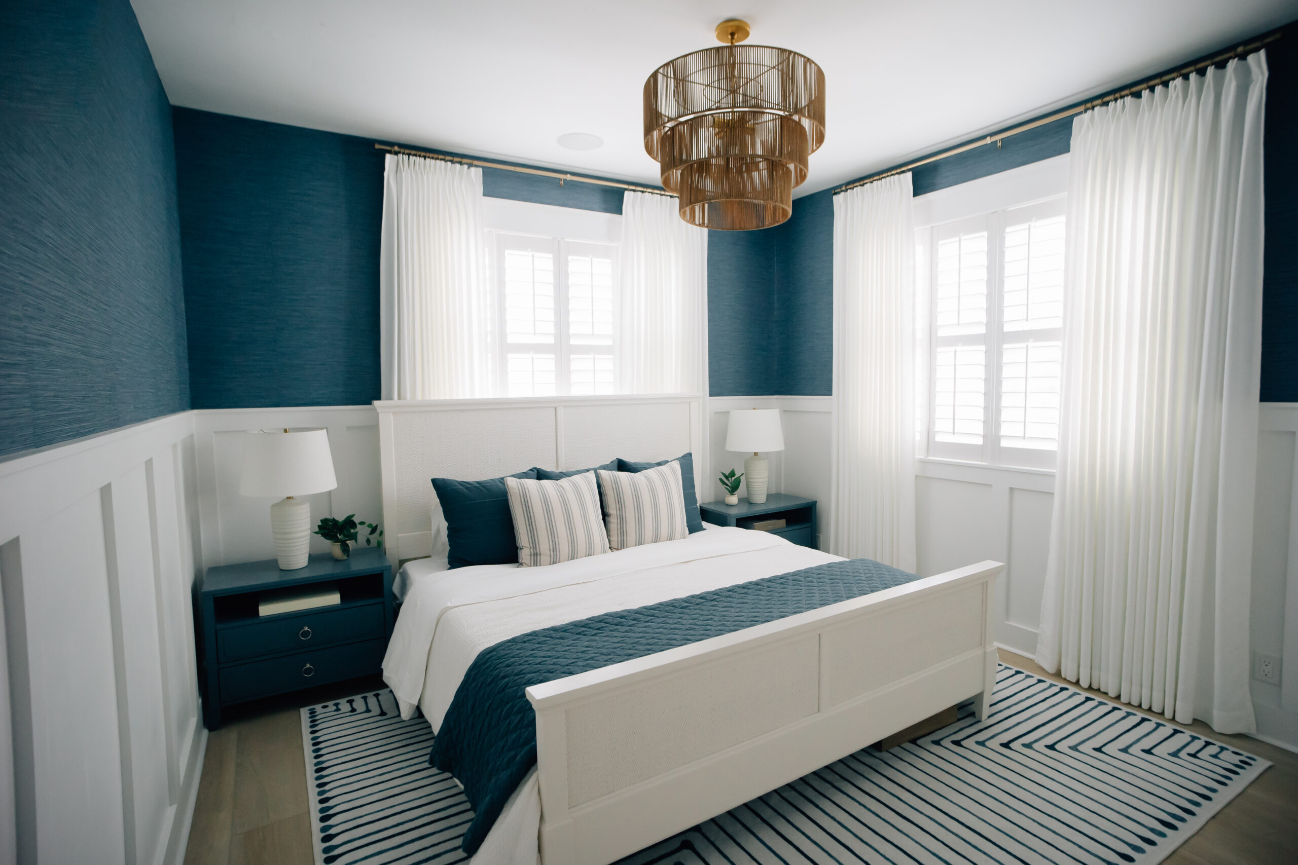 King bed with natural-wood head and baseboards in front of floor-to-ceiling curtains with modern nightstands and elegant table lamps.